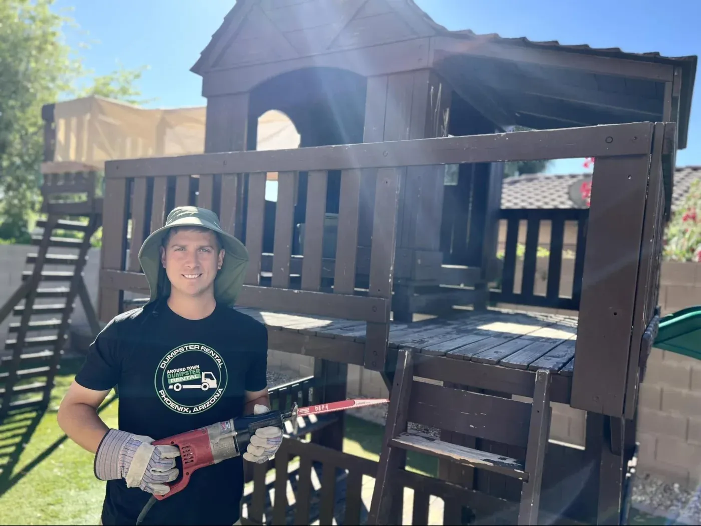 A crew member with a reciprocating saw beside a wooden playset being dismantled in Surprise